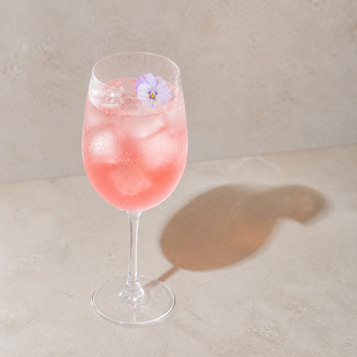 Date Night light pale-red spritz served over ice in a stemmed wine glass on warm stone, garnished with an edible viola resting inside the glass